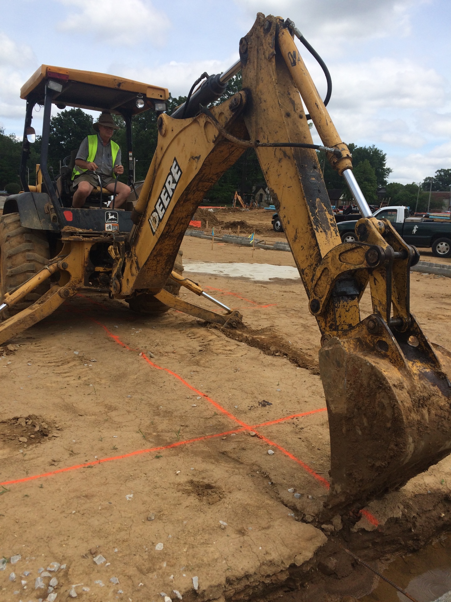 A construction worker wearing a high-visibility safety vest operating a yellow John Deere backhoe excavator to dig a trench at a dirt construction site, following bright orange spray-painted survey lines on the ground.