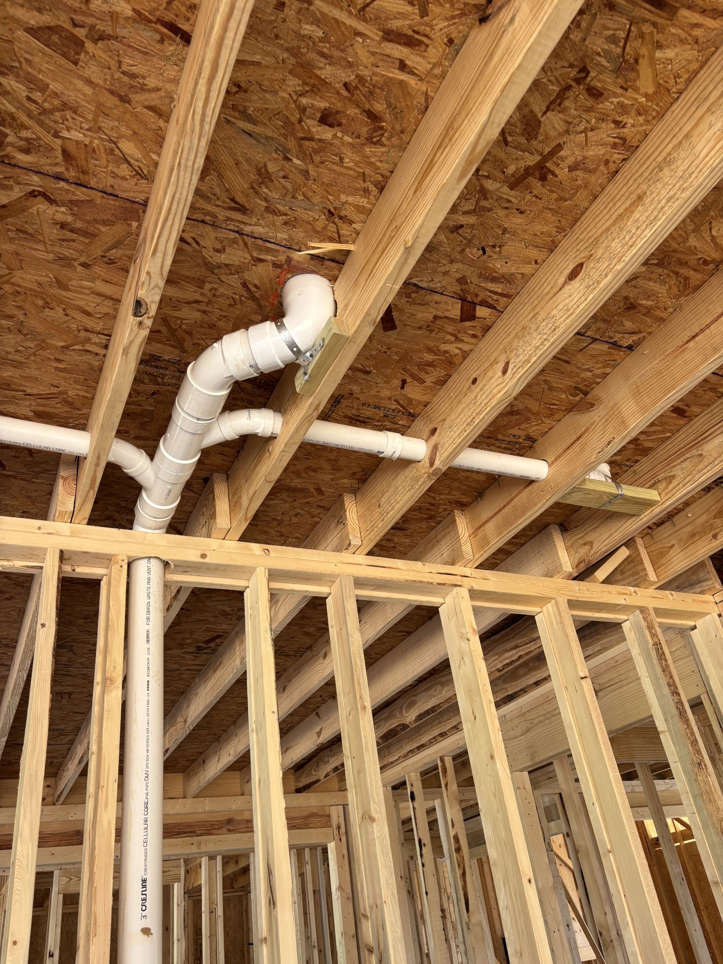 Low-angle view of a residential rough-in plumbing installation inside an open wooden framing structure, showing white PVC drain and waste pipes running through and supported beneath the ceiling joists.