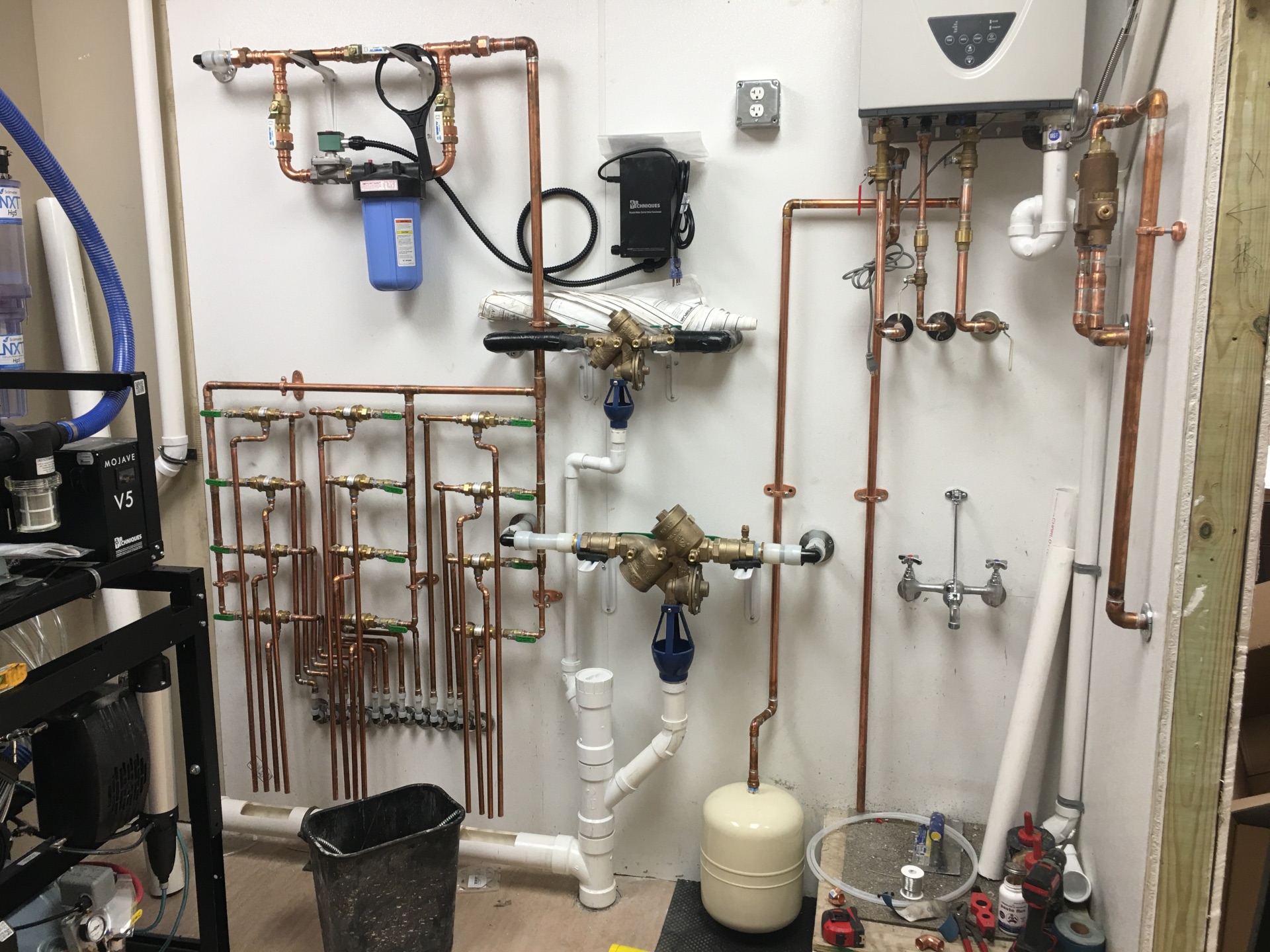 A mechanical wall installation showing a complex plumbing assembly with organized copper piping lines, a blue water filtration housing, a small cream-colored expansion tank, white PVC drain lines, and electrical control units mounted to a wall.