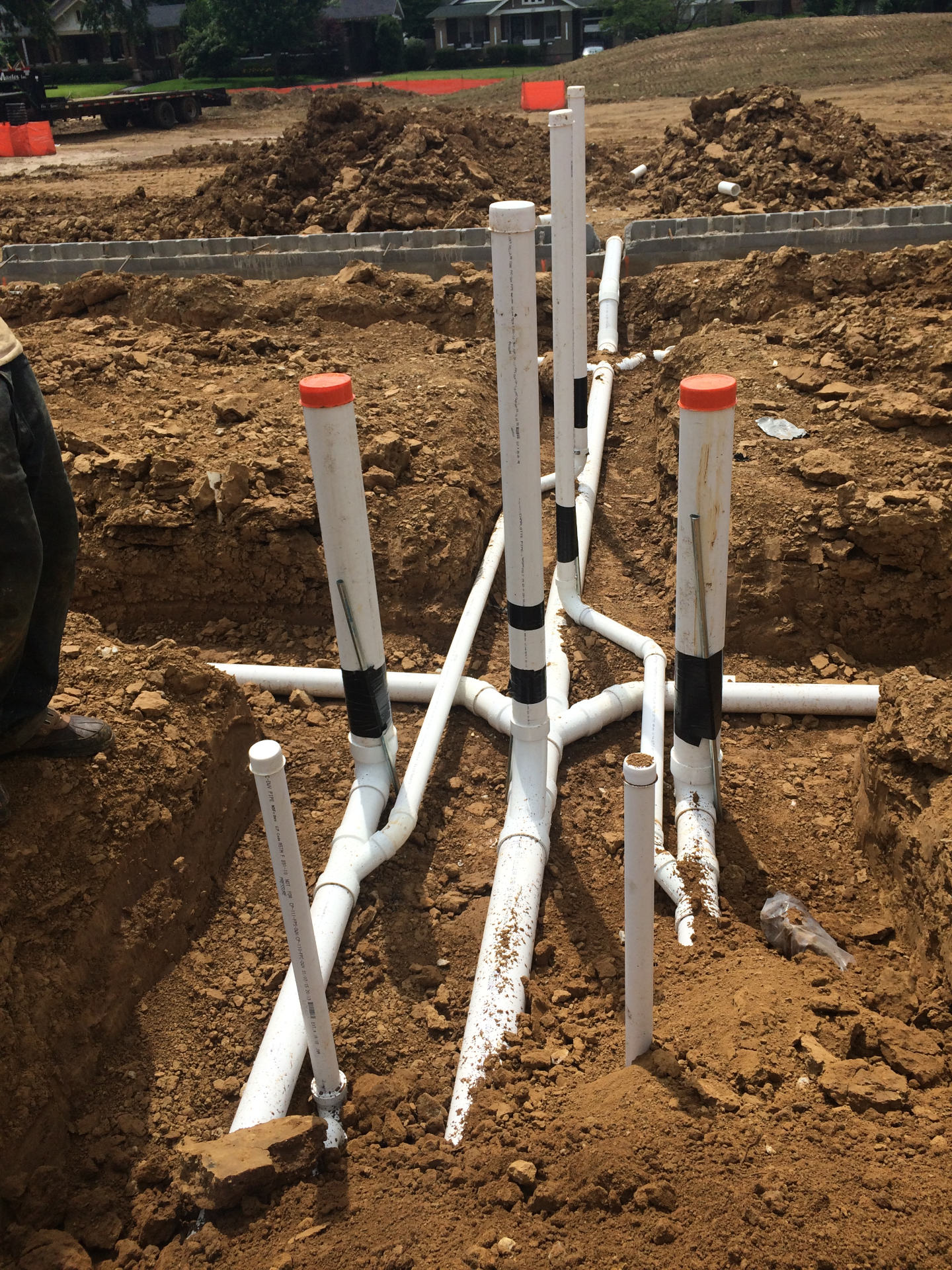 An underground plumbing installation at a construction site, showing a network of white PVC pipes, connectors, and vertical risers routed through dug-out dirt trenches."