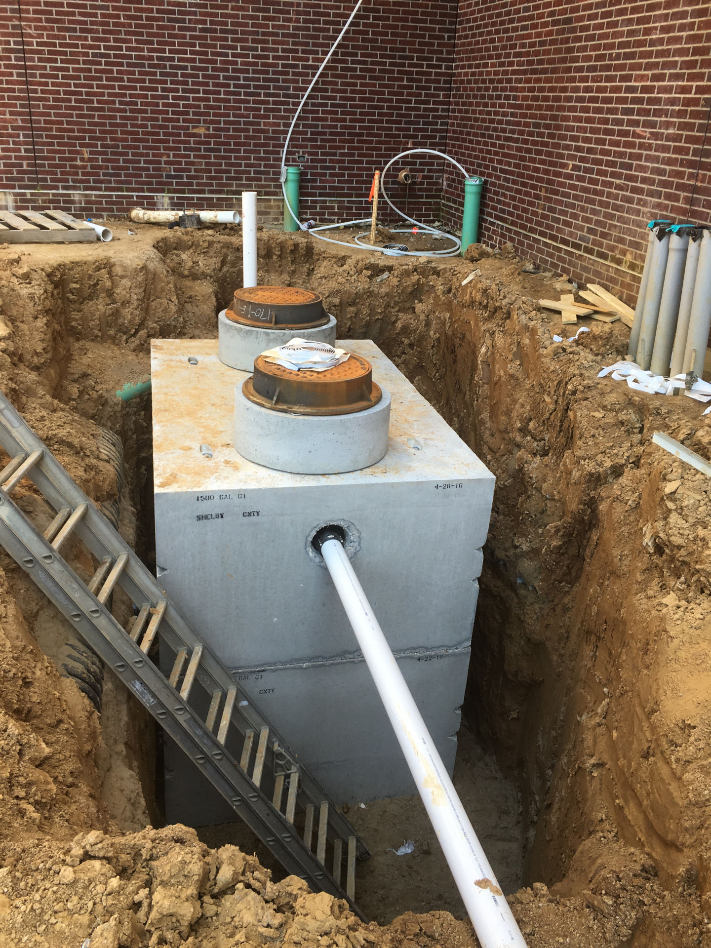 A large concrete grease trap or septic tank being installed in a deep dirt excavation pit next to a brick building, with connected PVC plumbing pipes and an aluminum ladder leaning against the side.