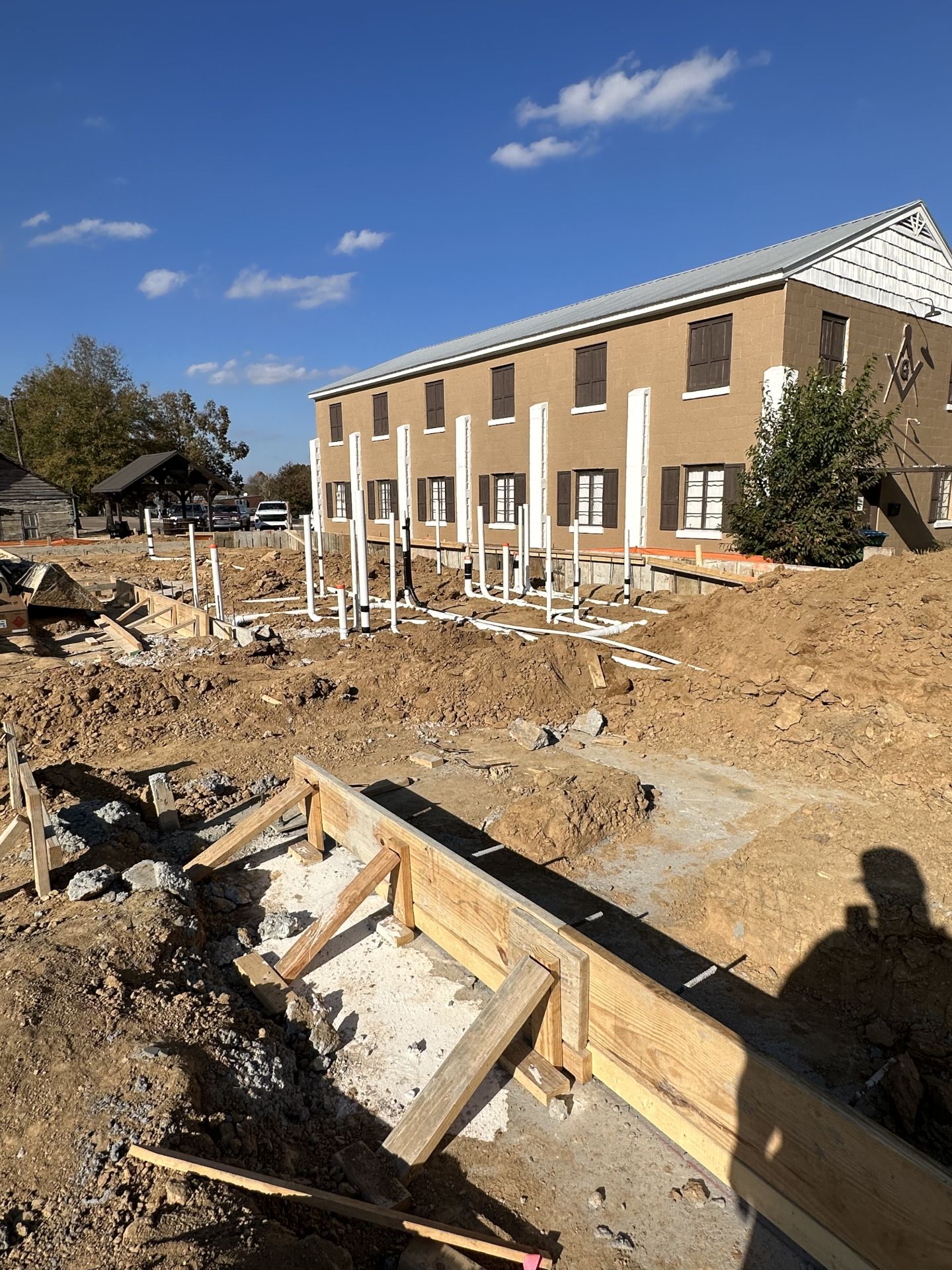 An active outdoor construction site showing wooden foundation formwork and a grid of white PVC plumbing pipes rising from the dirt, set in front of a two-story tan building under a clear blue sky.