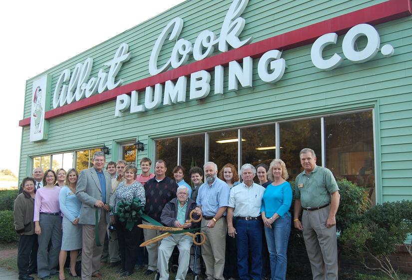 "A large group of people gathered outside the Albert Cook Plumbing Co. office building for a ribbon-cutting ceremony, with two men in the center holding giant wooden ceremonial scissors."