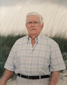 A medium shot portrait of an elderly man with short white hair and large aviator-style glasses, looking directly at the camera. He is wearing a short-sleeved, light blue and white plaid button-down shirt tucked into light-colored trousers with a dark leather belt. The background features tall beach grass or dunes under an overcast, hazy sky.